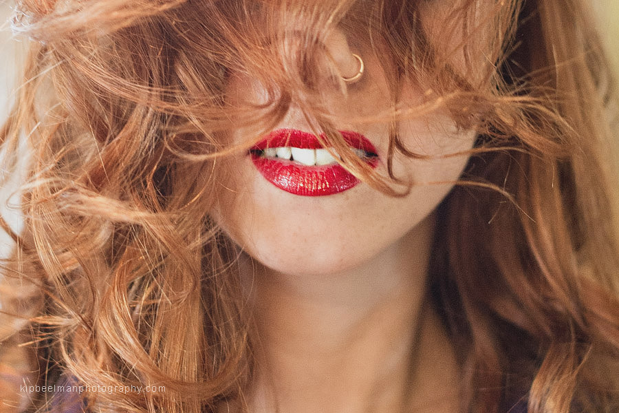 A bride with red hair has her long hair strewn over her face during hair styling from her Golden Gardens Bathhouse wedding to reveal only her nose ring and lips with bold red lipstick