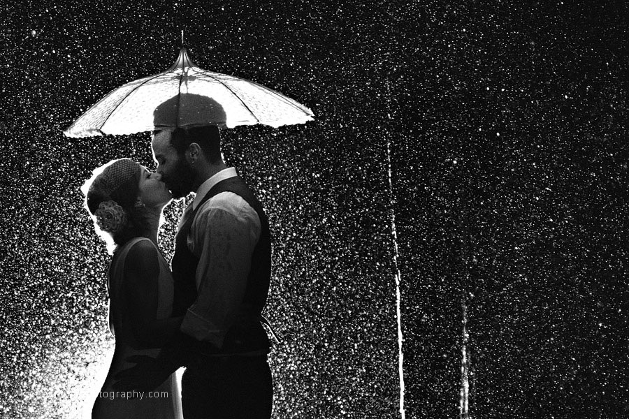 A Golden Gardens Bathhouse wedding couple are photographed under a vintage parasol from Bella Umbrella at night with a backlight to highlight the torrent of rain falling around them