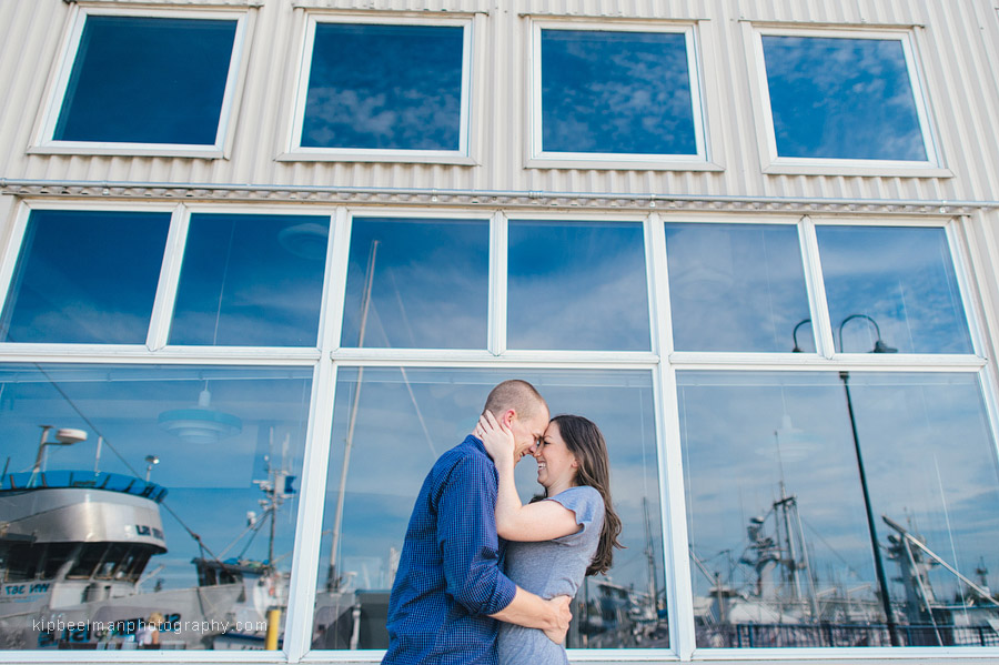 A smiling couple pose with their heads together in embrace in front of a building reflecting blue sky and boats in the windows during their Fishermens Terminal engagement session